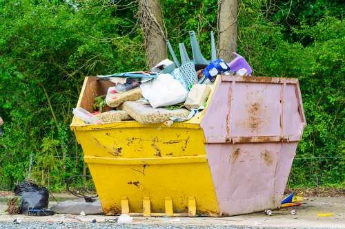 A large, rectangular skip with a yellow base and pinkish-bink upper section, positioned on a gravel surface outdoors, filled with miscellaneous household rubbish including plastic bags, cardboard boxes, a white plastic container, and various other debris. The skip is situated near a backdrop of lush green trees and bushes, with some black plastic trash bags placed on the ground nearby. The scene indicates a waste collection or clearance activity consistent with commercial or domestic rubbish removal services, as seen in areas serviced by companies like Office Clearance Osterley focusing on rubbish disposal within the Osterley or surrounding London postcode regions.