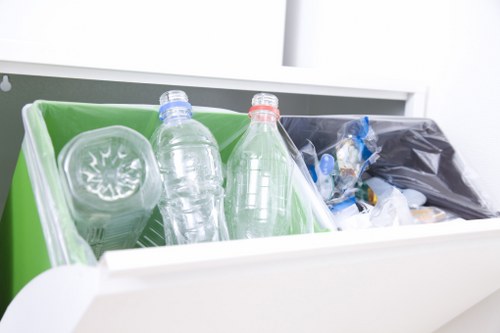 Close-up of a partially open white waste disposal drawer containing a green plastic bin with a clear plastic bottle and a red-capped bottle, alongside a black rubbish bag filled with miscellaneous waste. The scene suggests a domestic setting, possibly in an office environment or kitchen area, with clean surroundings and natural lighting reflecting off the plastic bottles. The presence of recyclable bottles and general waste indicates an organized approach to rubbish disposal, consistent with rubbish removal services offered by Office Clearance Osterley. The image highlights materials such as transparent, smooth plastic bottles and the contrasting black rubbish bag, emphasizing waste separation and collection efforts typical in commercial or residential cleanouts in the Osterley area.