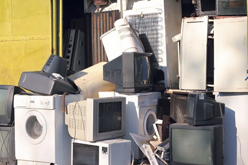 A collection of discarded electronic waste and household appliances is piled outdoors, including several CRT and flat-screen computer monitors, washing machines, and a microwave oven. The items are stacked haphazardly, with some objects leaning against each other and others resting on the ground or atop nearby machinery. The appliances exhibit various finishes, such as white and black plastic casings, with some showing signs of dirt and weathering. In the background, a yellow building or container with a textured surface is partially visible. The overall environment appears to be an outdoor waste collection or recycling yard, consistent with rubbish removal services offered by companies like Office Clearance Osterley, serving suburban areas such as Osterley or nearby towns. The scene suggests a focus on electronic and appliance disposal, with the appliances occupying a significant portion of the space, ready for collection or recycling as part of a commercial or residential clearance project, typical of rubbish removal operations in the London borough or surrounding areas.