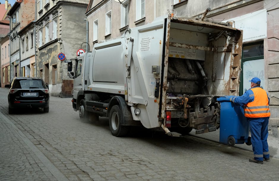 A middle-aged woman with short, dark hair is seen standing outdoors on a paved area, disposing of waste into a tall, round, stainless steel public rubbish bin with a slanted opening. She is dressed in