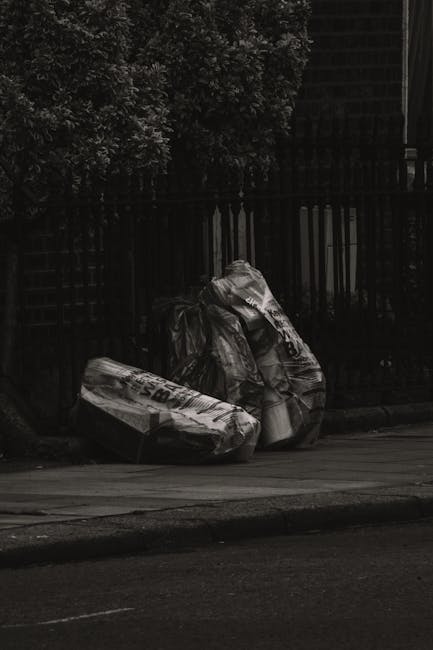 Several large black garbage bags filled with waste material are placed on the pavement beside a black metal fence and a tree with dark foliage. The waste bags appear full and somewhat crumpled, with s