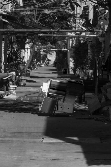 In the foreground, a person is engaged in a rubbish removal task, bending over and placing or sorting waste into a large, worn fabric sack that appears filled with various debris. Nearby, there are ca