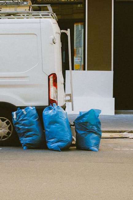 A blue plastic waste bin with a closed lid standing beside the back of a white delivery truck parked on a concrete surface. The bin appears to be made of durable, slightly textured plastic with some v