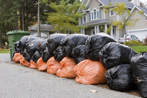 A row of black and orange plastic rubbish bags filled with waste, stacked along the edge of a residential street, with some bags leaning slightly and others tightly packed. The bags are placed on the pavement in front of a green wheelie bin, with the black bags appearing to contain general rubbish and the orange bags possibly indicating a specific waste type or recycling collection. In the background, there are leafy trees, a driveway, and a two-storey detached house with white exterior walls, light grey roofing, and multiple windows. The scene takes place during daylight, with natural light illuminating the bags and surroundings, suggesting an organised clearance or waste collection process typical of UK residential areas. This imagery exemplifies the rubbish removal services provided by Office Clearance Osterley, supporting efficient waste disposal in the Osterley region.