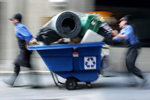 A blue recycling bin mounted on nearby wheels is being pushed along a street by two individuals in blue uniforms and black hats, both blurred to indicate motion. The bin is filled with various waste items, including a large black garbage bag, a green wheeled bin, a rolled-up black object, and a white plastic container, all visibly overflowing from the top of the bin. The background shows a concrete building facade and a roadway with moving vehicles, emphasizing an urban setting. The scene suggests waste collection or rubbish removal activity typical of a professional service operating in a busy town environment, possibly in Osterley or surrounding areas, with the focus on efficient rubbish disposal and recycling efforts from a local rubbish removal company such as Office Clearance Osterley.