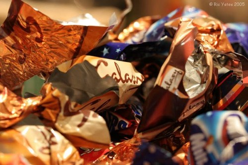 A close-up image of a pile of crumpled foil wrappers in various colors including metallic copper, silver, gold, and vibrant patterned designs, with some wrappers displaying partially visible text and logos. The wrappers are stacked haphazardly, filling the frame and creating a textured surface with reflections and creases. The background is blurred and indeterminate, emphasizing the shiny, reflective surfaces of the foil. The composition suggests waste accumulation, typical of rubbish that might be collected by a professional rubbish removal service in Osterley or nearby areas, as provided by Office Clearance Osterley, in a setting that could be an indoor or outdoor environment such as a kitchen or a waste collection point.